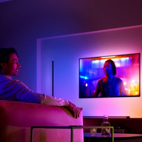 Televisor montado en la pared con luces que se sincronizan con el contenido en su pantalla mientras un hombre mira desde el sofá. 