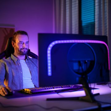 Un hombre sentado frente a un monitor de PC equipado con una tira de luz Hue Play gradient lightstrip que brilla en tonos de luz azul y púrpura. 