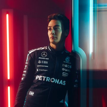 El piloto de F1 George Russell se sienta en un sofá beige en una sala de estar con una iluminación suave y cálida.