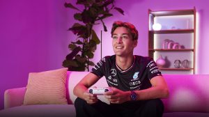 El piloto de F1 George Russell está sentado en un sofá con el mando a distancia de un videojuego en una habitación con iluminación rosa.