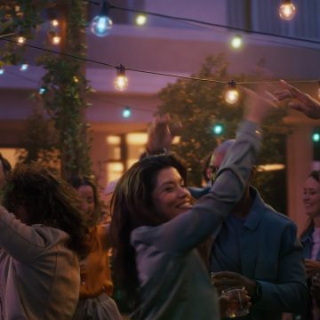 Une femme danse sur une terrasse, sous des guirlandes lumineuses extérieures à globes Festavia qui diffusent une lumière connectée bleue, rose et blanche.