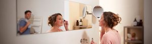Une personne se maquille devant un miroir de salle de bain baigné d’un éclairage vif et précis, idéal pour des soins méticuleux, avec en arrière-plan des flacons et des étagères bien organisés