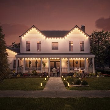 De gevel, daklijn en veranda van een huis verlicht met Festavia permanente buitenlampen die in helder wit licht schijnen.