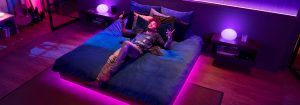 Persoon die naar muziek luistert op een bed in een slaapkamer, omringd door kleurrijke LED-lampjes die synchroon met de muziek knipperen in de tinten roze, paars en blauw.