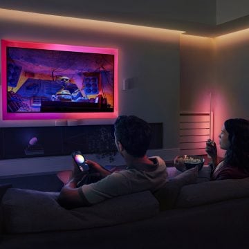 Een stel zit op een bank en kijkt tv. Ze worden omgeven door indirect licht in roze tinten van een Hue Flux lightstrip. De man bedient de lampen met de Hue app op zijn smartphone. 