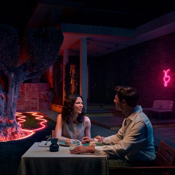 Et par spiser på terrassen med neonlys som omkranser et tre og et blomsterbed og lyser i rosa fargetoner. 