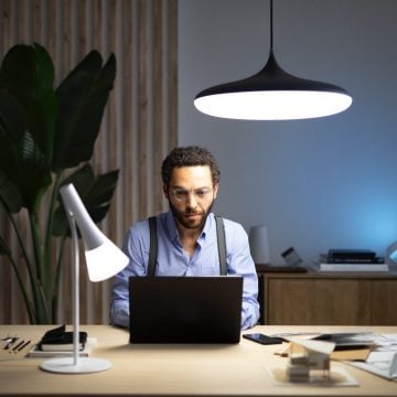 Mężczyzna siedzi przy laptopie ustawionym na biurku i oświetlonym inteligentną lampą stołową i lampą wiszącą, które dają chłodne, białe światło.